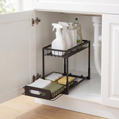 A black metal two-tier under-sink cabinet organizer with a pull-out bottom tray, shown installed inside an open white bathroom vanity. The organizer holds white spray bottles, a green bottle, folded towels, and cleaning sponges.
