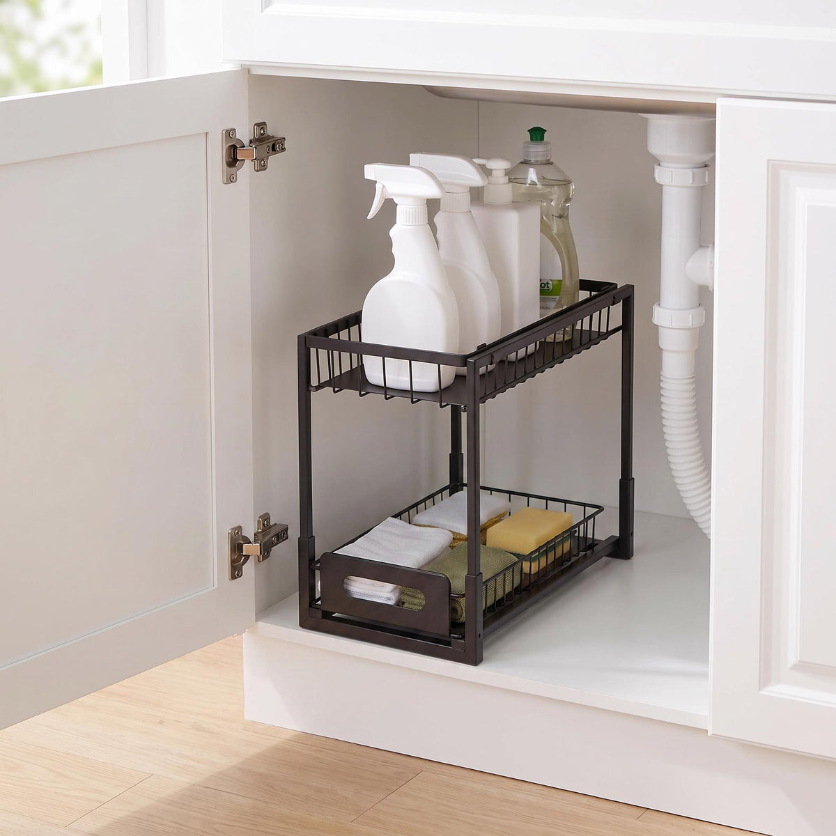 A black metal two-tier pull-out organizer basket inside a white kitchen cabinet, holding cleaning spray bottles and sponges.