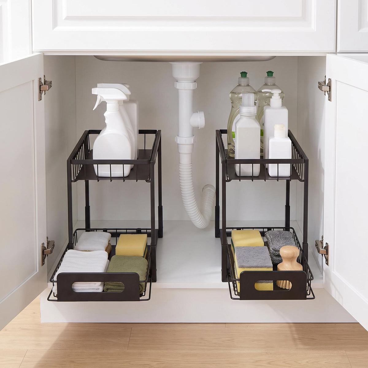 Two black metal under-sink cabinet organizer baskets shown inside an open white bathroom or utility cabinet, holding cleaning supplies, towels, and sponges.