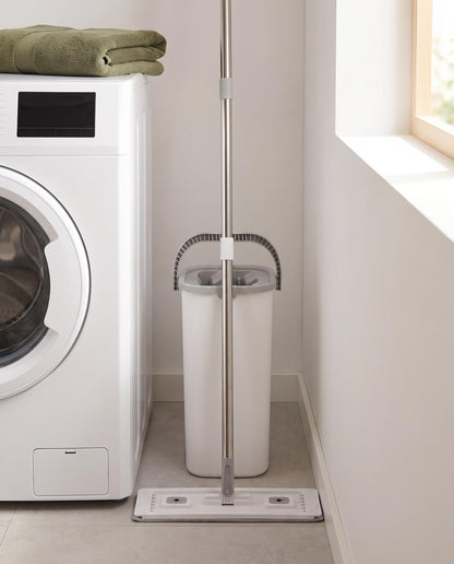 Flat mop and narrow dual-chamber bucket stored upright in the gap beside a white washing machine