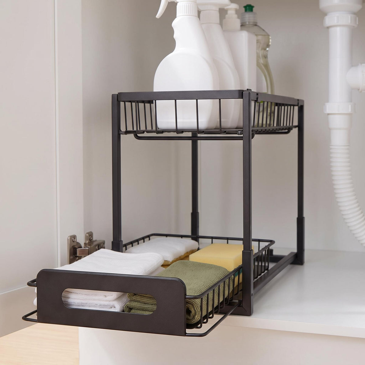 A two-tier black metal under-cabinet organizer shelf with a pull-out drawer, shown holding cleaning spray bottles on the top rack and folded towels and a sponge on the lower rack.