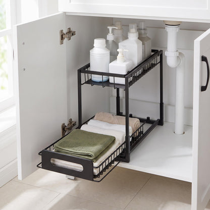 A black metal two-tier under-sink storage organizer with a pull-out bottom tray, shown inside a white bathroom cabinet containing toiletries and folded towels.